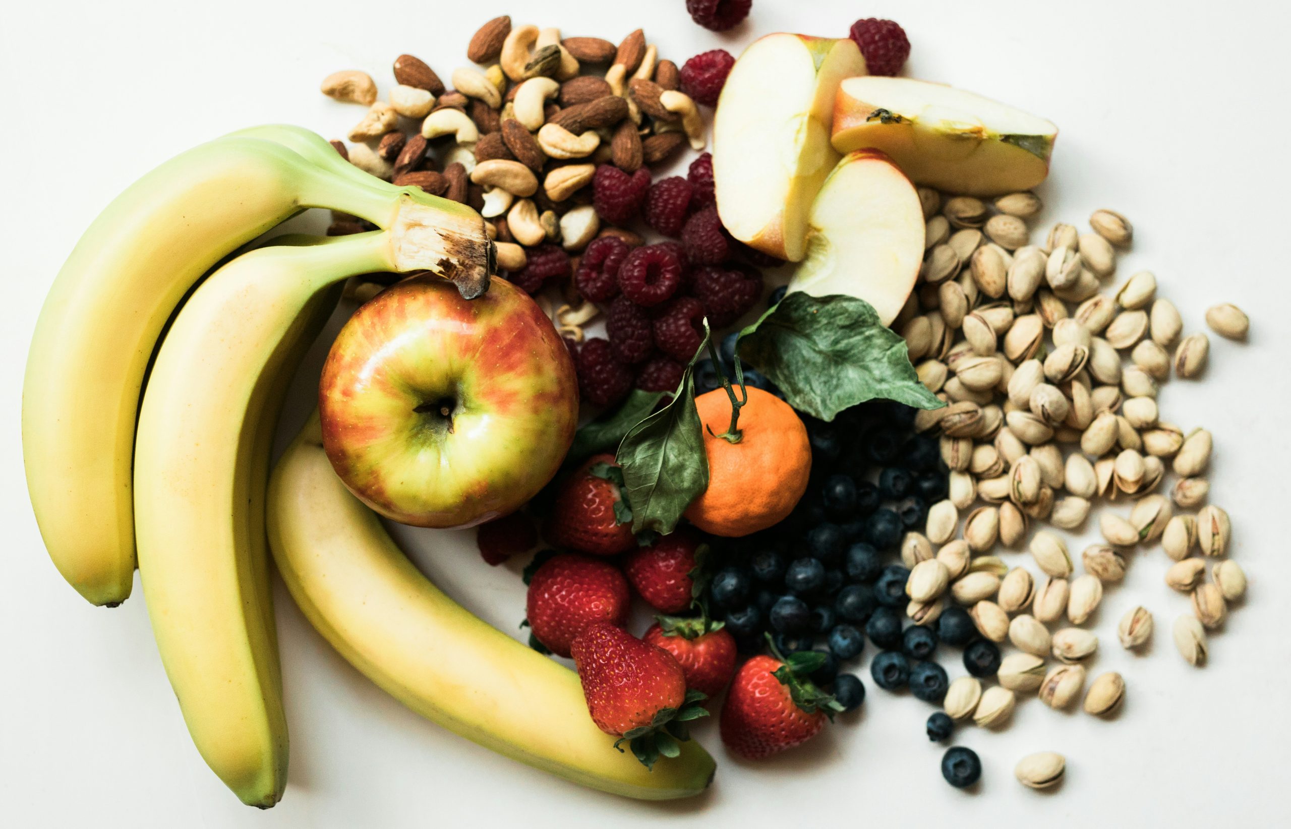 Assorted fruits including bananas, apple, orange, strawberries, blueberries, raspberries, and nuts such as almonds and pistachios arranged on a white surface.