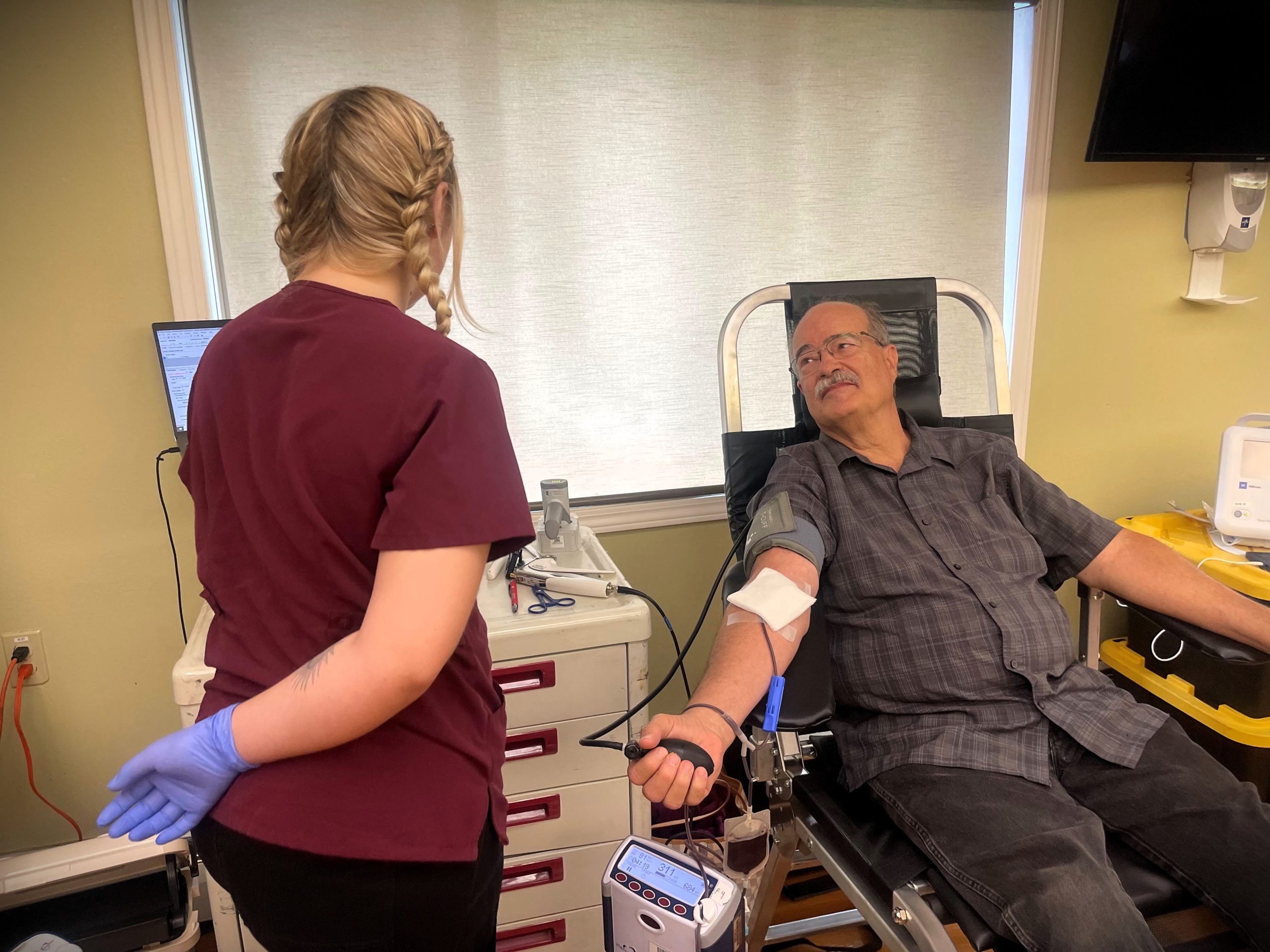 A man donates blood while seated, holding a stress ball, as a healthcare worker in scrubs and gloves stands nearby in a medical room.