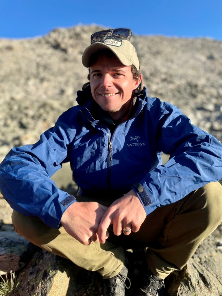 A person wearing a blue jacket, tan cap, and khaki pants sits cross-legged on rocky terrain outdoors with sunlight casting shadows.