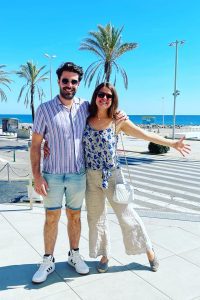 A man and a woman stand smiling together outdoors near a beach, palm trees, and a pedestrian crossing on a sunny day.