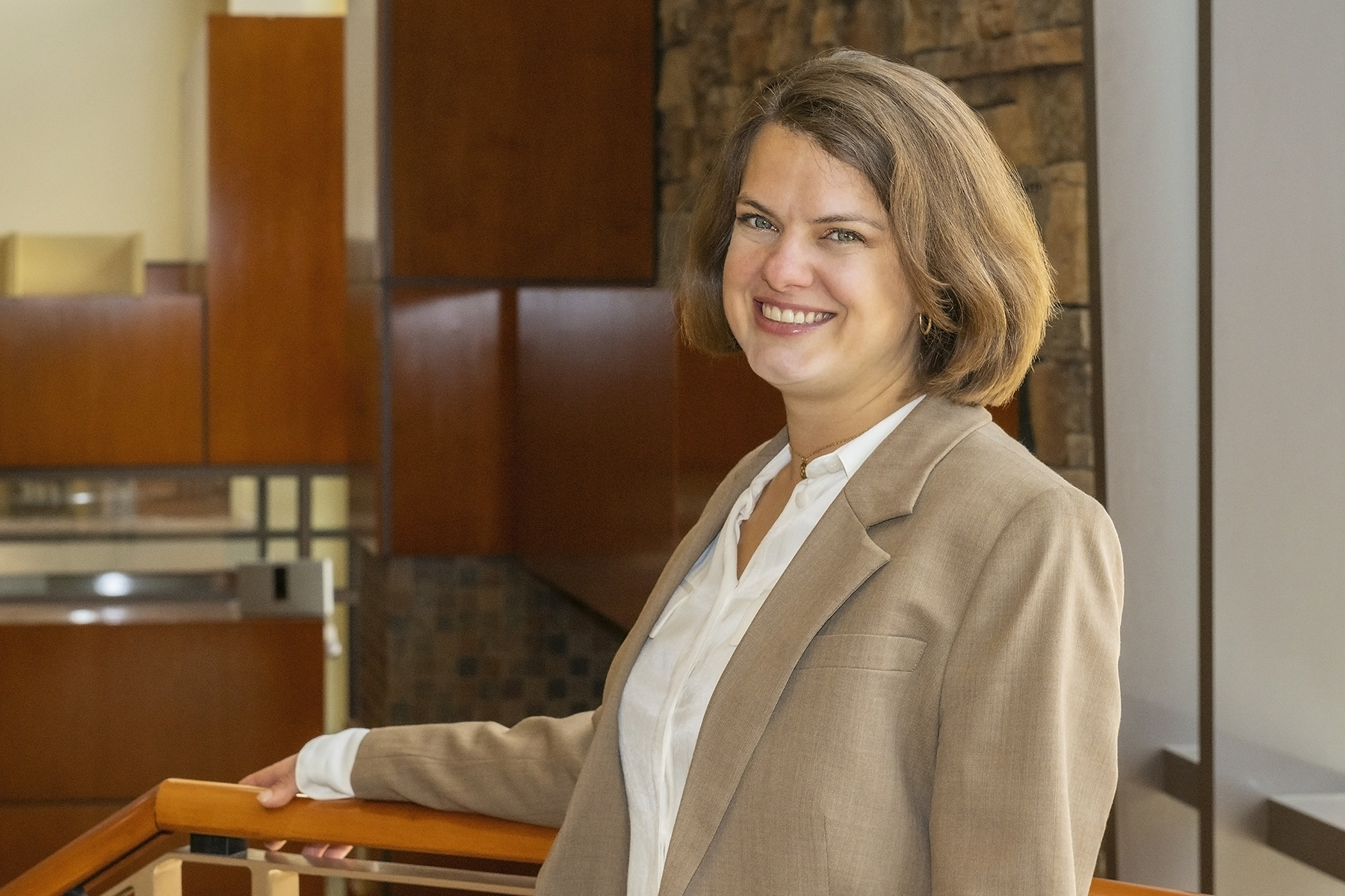 A woman in a tan blazer and white shirt stands indoors, smiling and resting her arm on a wooden railing with a modern interior background.