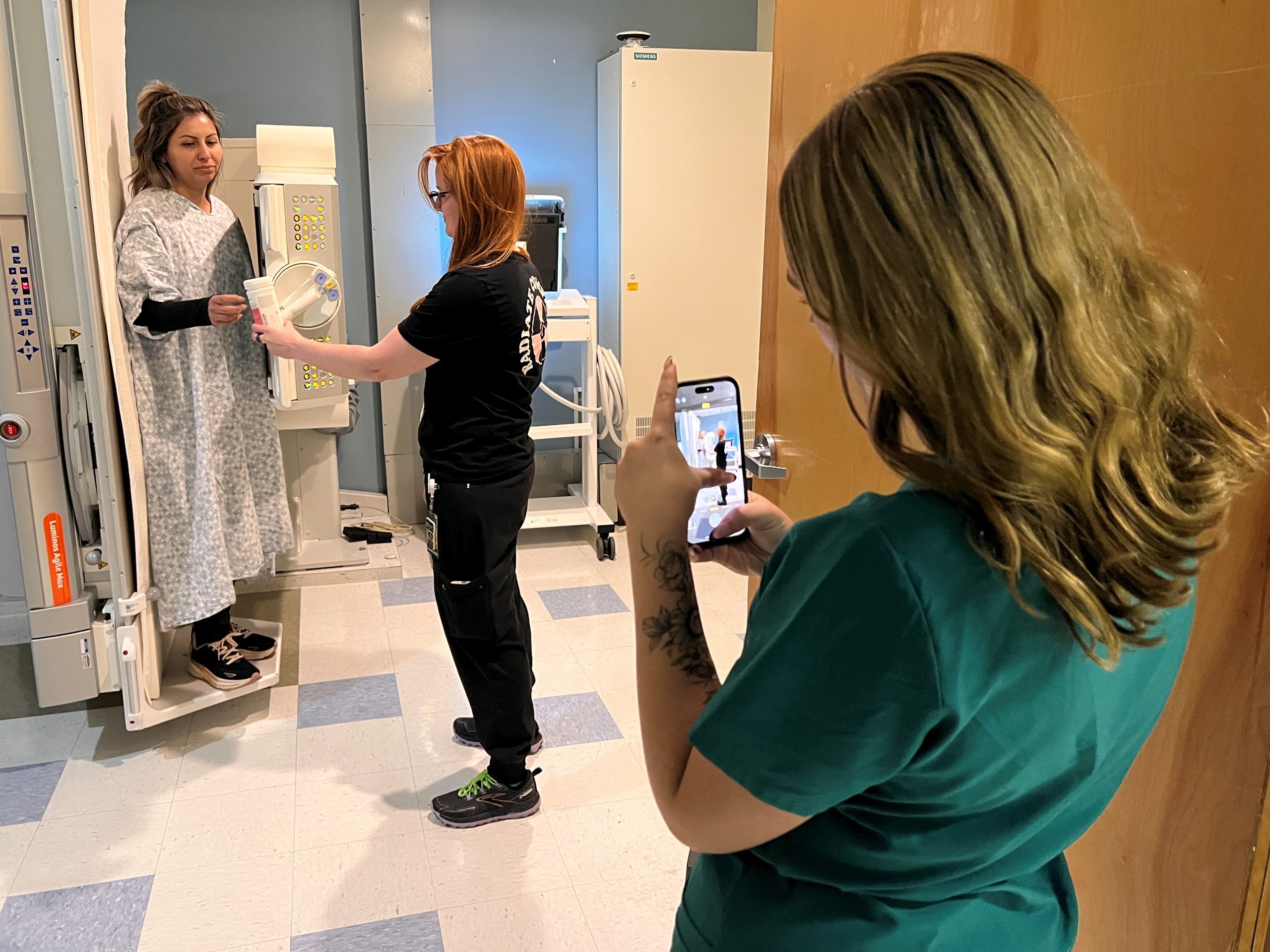 A woman in a hospital gown stands by an X-ray machine, while one person assists her and another takes a photo with a smartphone.