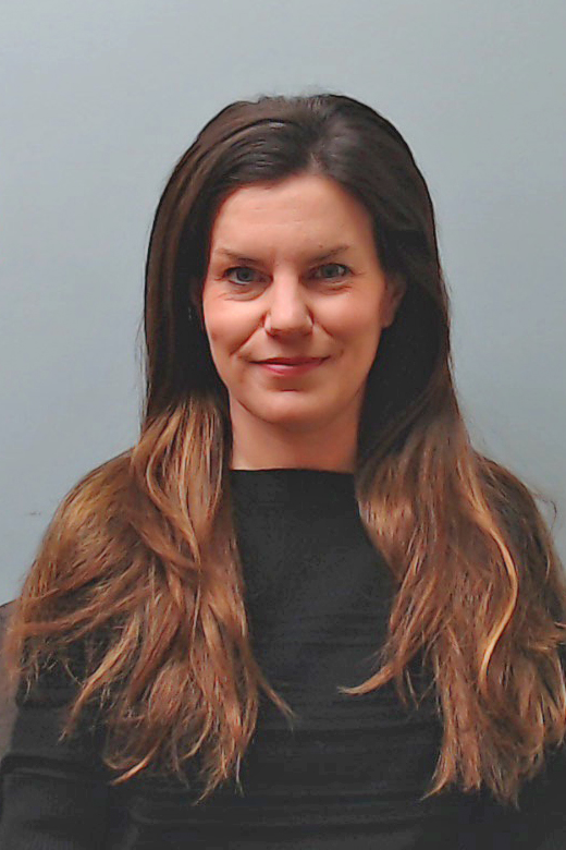 Woman with long brown hair wearing a black top, sitting against a plain light gray background, looking at the camera with a neutral expression.