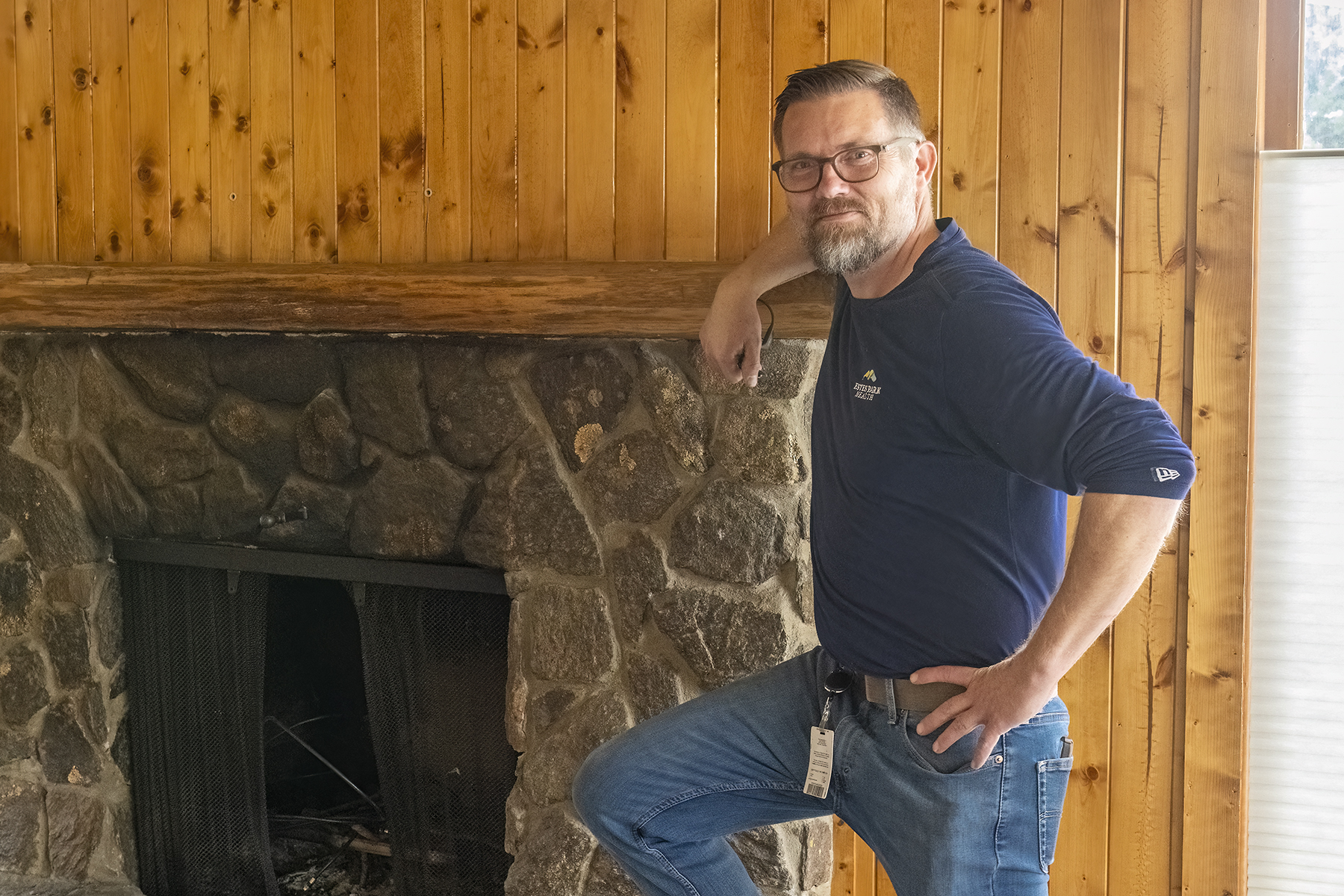A man with glasses and a beard stands next to a stone fireplace in a room with wood-paneled walls.