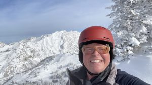 A person wearing a red helmet and goggles smiles for a selfie in a snowy mountain landscape with trees and peaks in the background.
