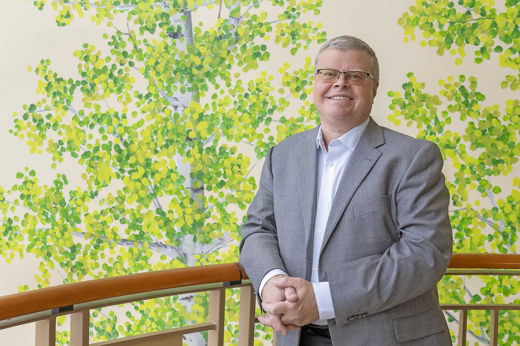 A man in a gray suit stands by a railing in front of a wall painted with green and yellow leafy trees.