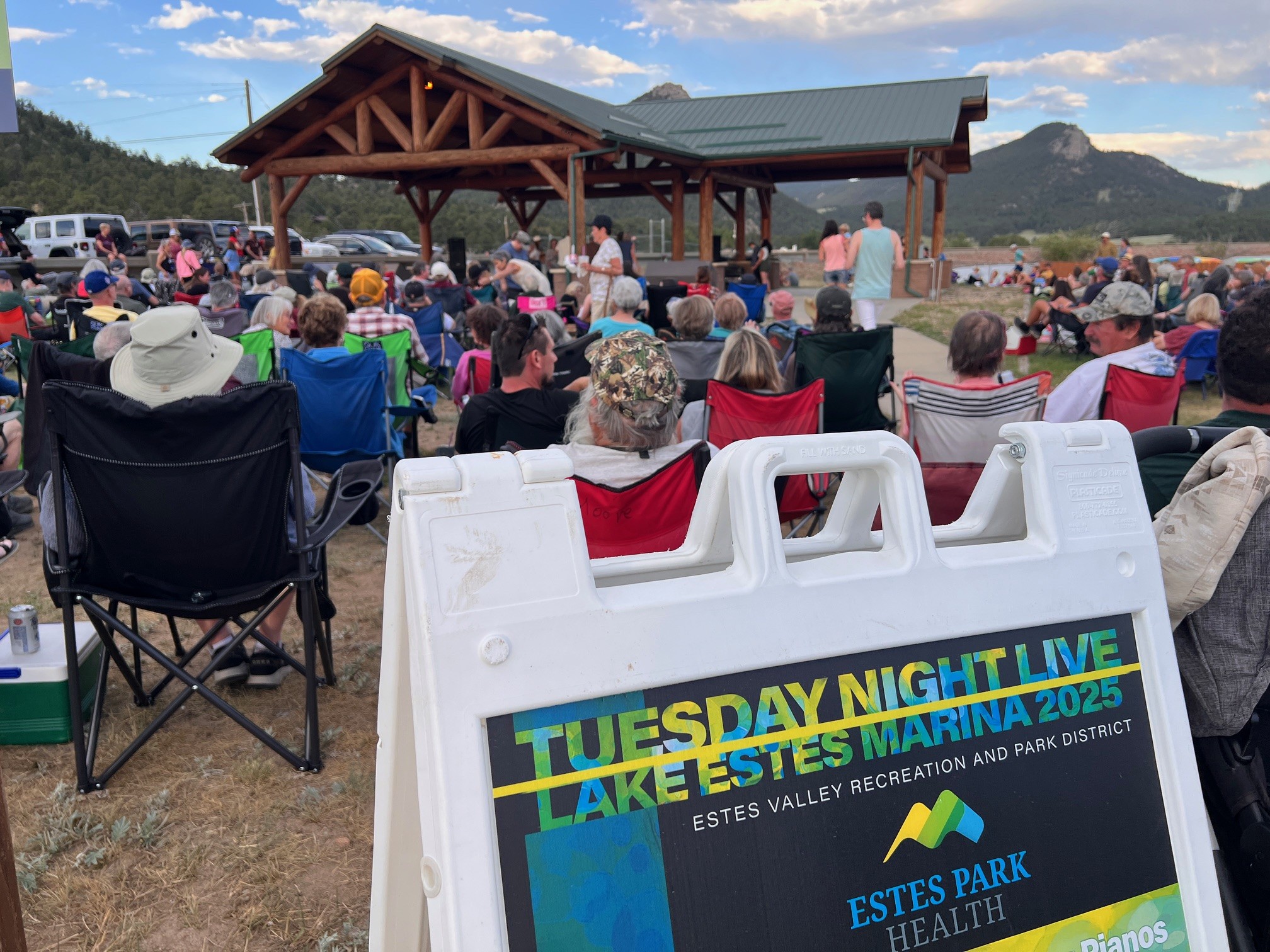 A crowd sits in chairs facing an outdoor stage at Lake Estes Marina for a Tuesday Night Live event, with a sign showing event details in the foreground.
