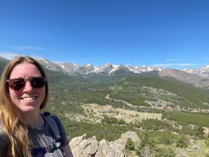 A woman wearing sunglasses and a backpack smiles at the camera with a scenic mountain range and forested valley in the background under a clear blue sky.