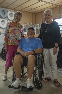 Two women stand beside a seated man in a wheelchair inside a room with shelves holding various items and stacked chairs in the background.