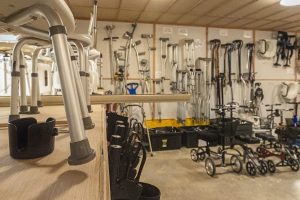 A storage room contains various mobility aids, including crutches, walkers, canes, wheelchairs, and shower seats, organized on shelves and against the wall.