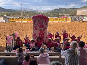 A group of women in pink shirts and cowboy hats pose in an arena with a large "Paint Estes Pink" sign during a rodeo event, while spectators watch from the stands.